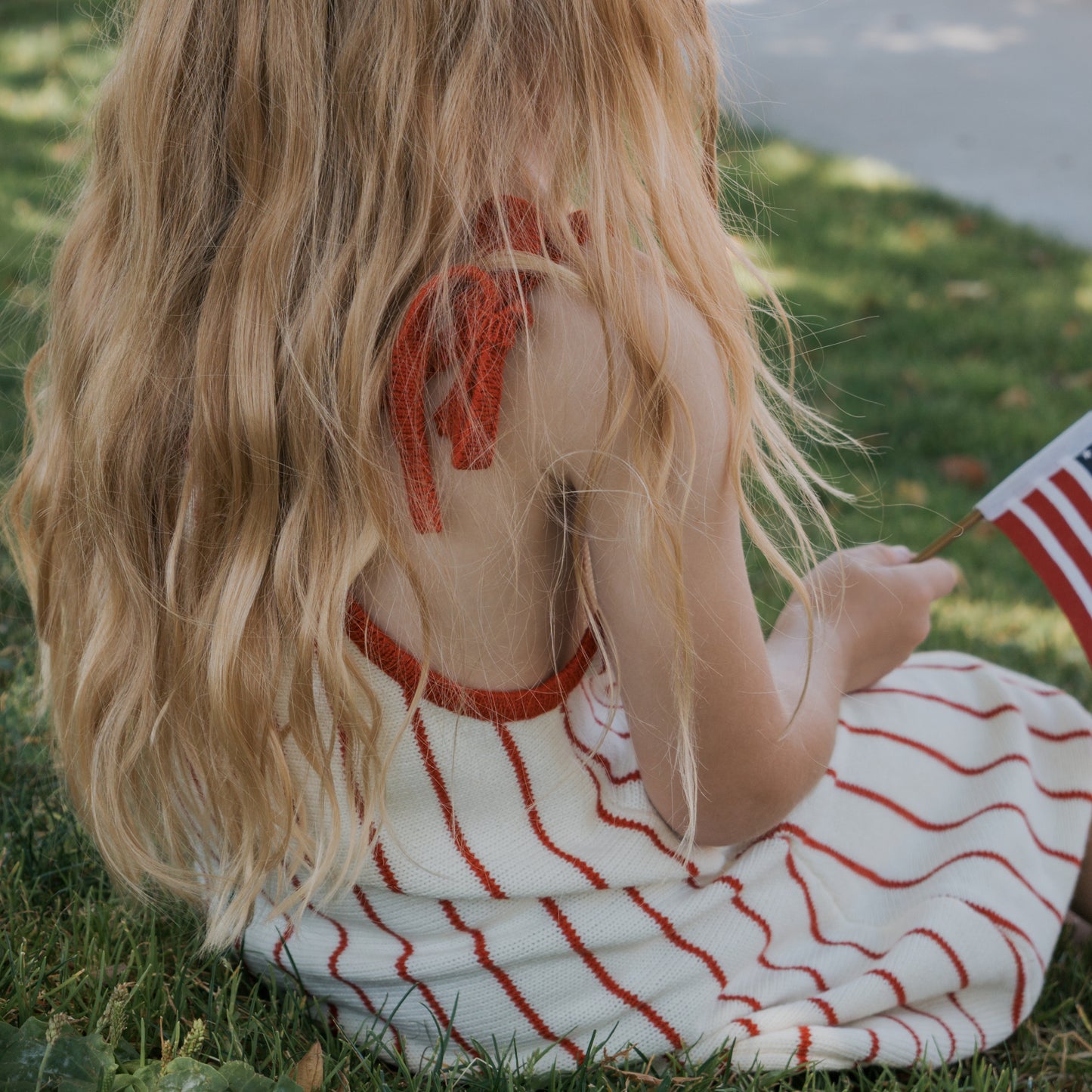 A young person with long, wavy blonde hair sits on the grass wearing the forever french baby Knit Dress | Picnic Pinstripe, a 100% cotton white dress with red stripes and adjustable tie straps, holding a small American flag. Their face is not visible.