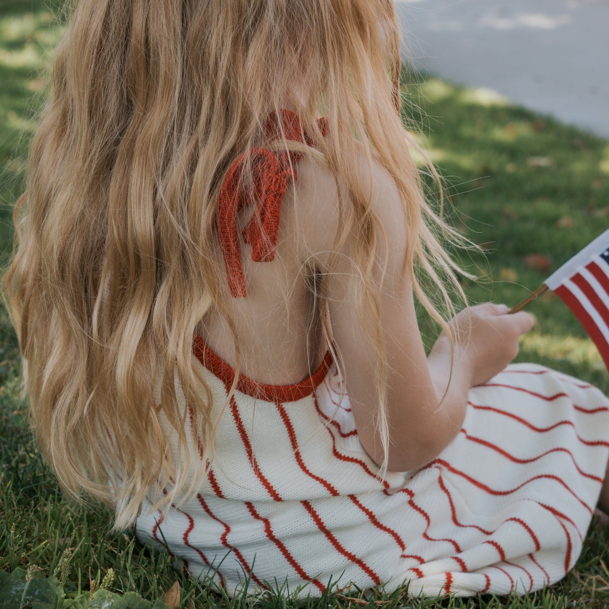 A young person with long, wavy blonde hair sits on the grass wearing the forever french baby Knit Dress | Picnic Pinstripe, a 100% cotton white dress with red stripes and adjustable tie straps, holding a small American flag. Their face is not visible.
