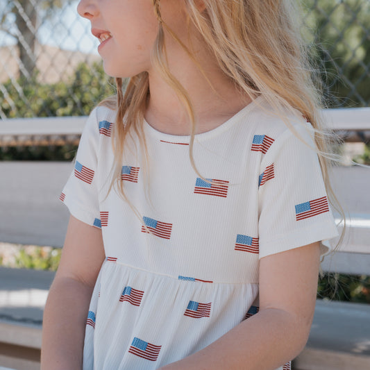 A young child with long blond hair wears the forever french baby Bamboo T-Shirt Dress | Americana Ribbed, sitting outdoors on a bench with a chain-link fence and trees in the background.