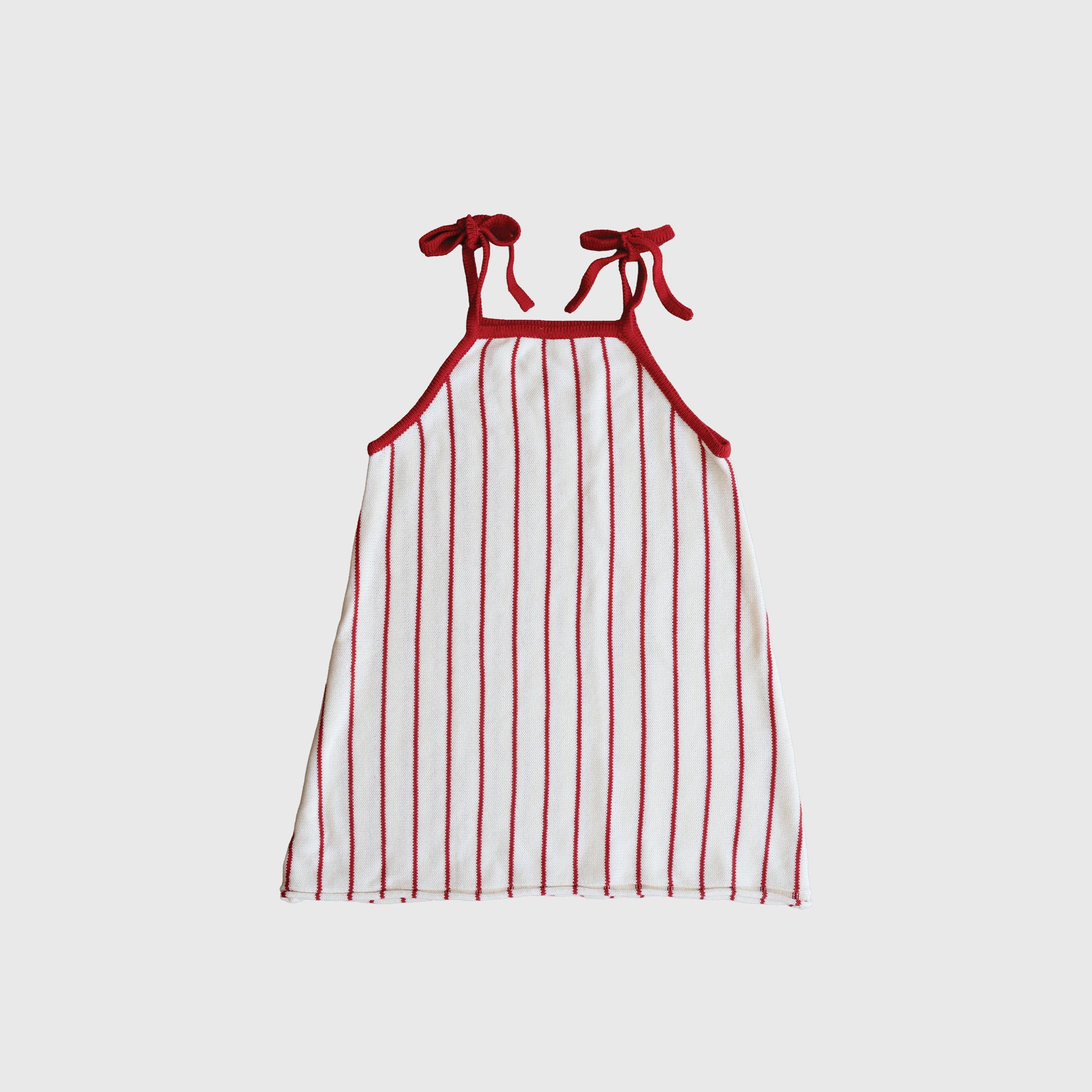 The Knit Dress | Picnic Pinstripe by forever french baby is a sleeveless white 100% cotton dress with vertical red stripes, red edging, and adjustable tie straps bowed at the shoulders, shown on a plain light gray background.