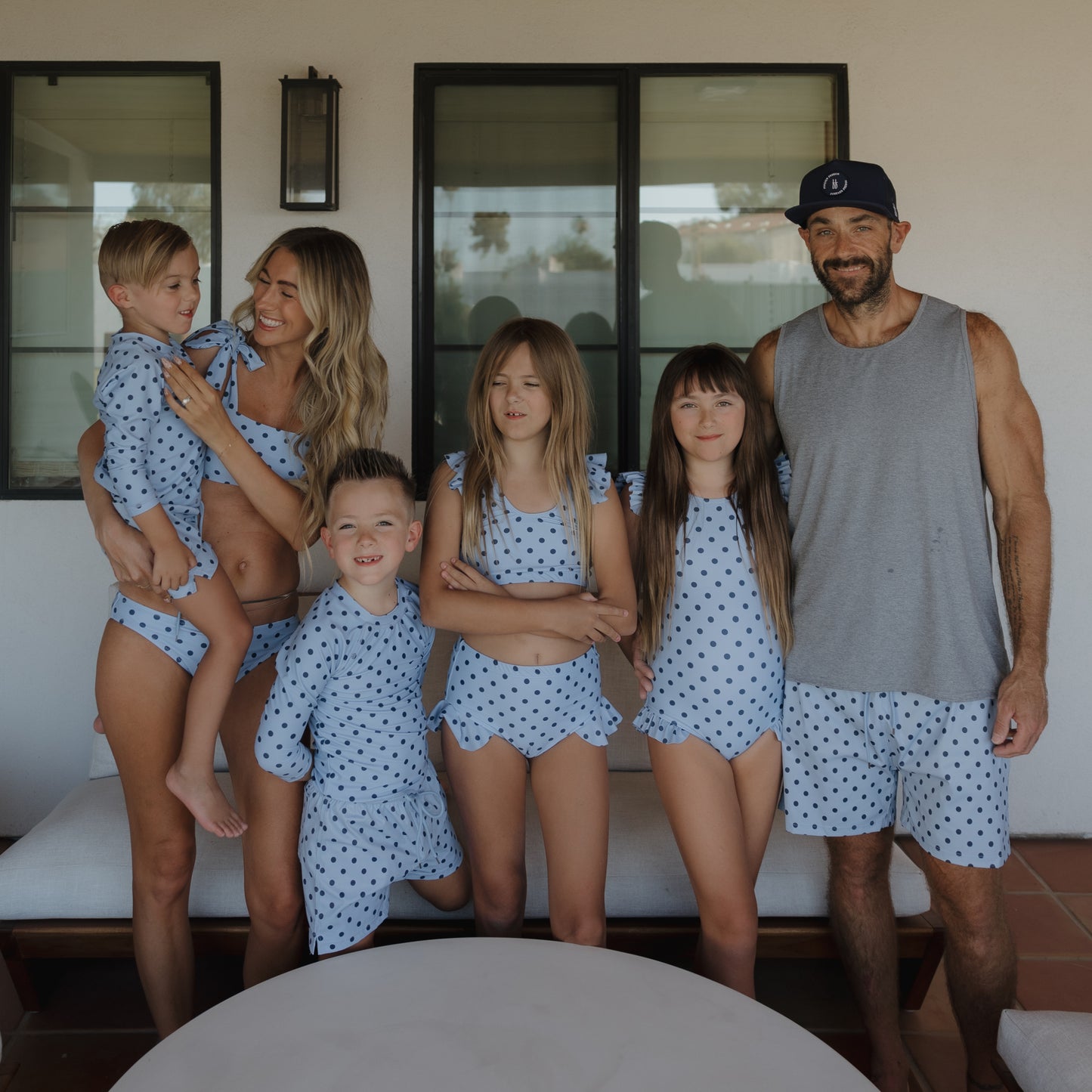 A smiling family of six poses outdoors by large windows and modern furniture, all dressed in forever French baby Girl's Sleeveless Swimsuit | Sully—matching blue and white polka dot one-pieces with ruffle shoulder straps for adults and kids.