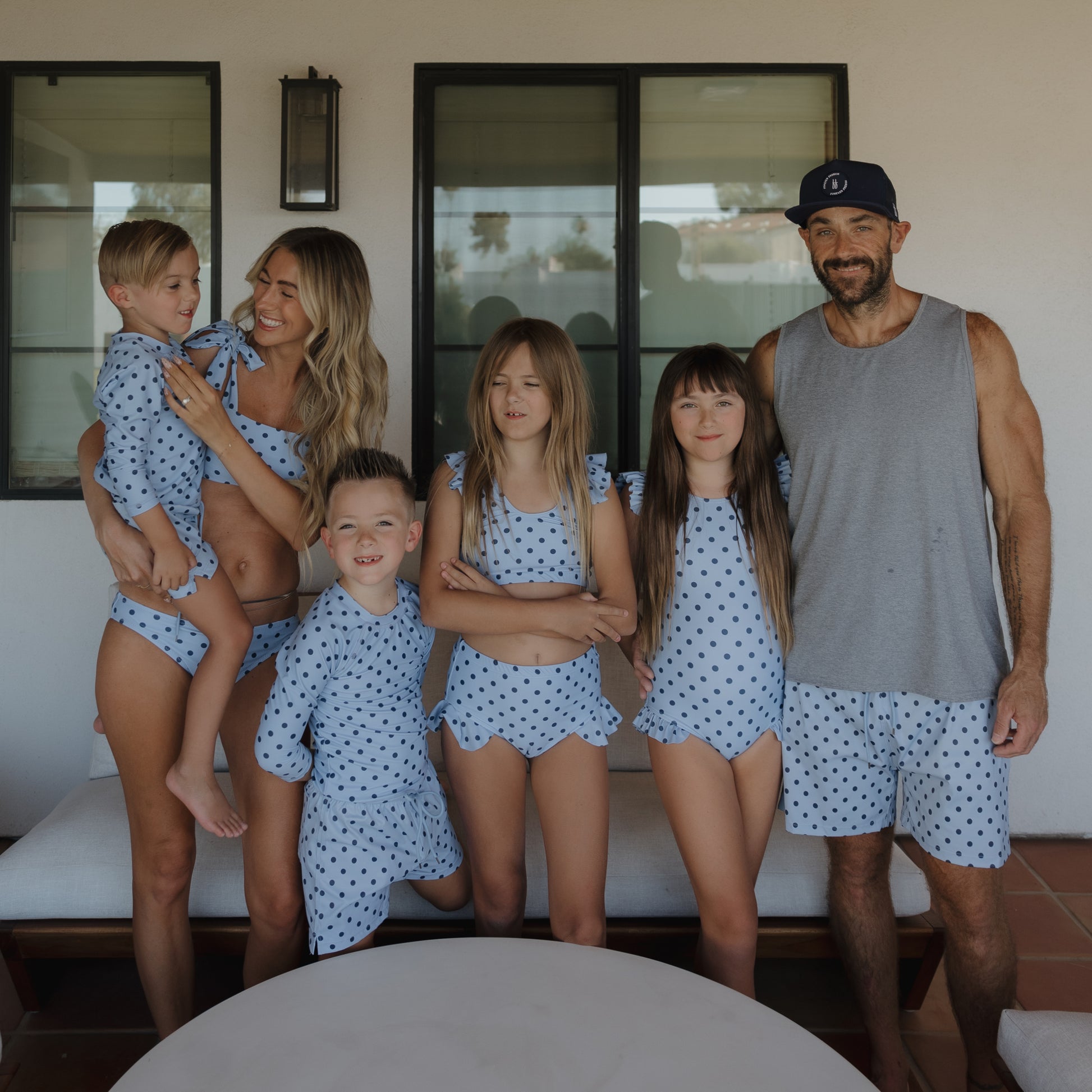 A smiling family of six stands outdoors in matching blue polka dot swimwear. The man wears forever french baby Men's Board Short | Sully and a tank top, while the rest wear coordinated polka dot swimsuits, two girls in the center, parents and two boys on each side.
