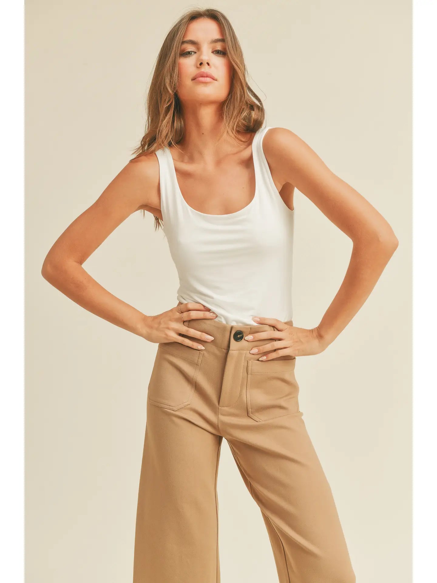 A woman with long wavy hair stands confidently with her hands on her hips. She is wearing the Double Layered Square Neck Tank Top in Off White from forever french baby and high-waisted beige pants. The background is plain light beige, giving a minimalist look to the image.