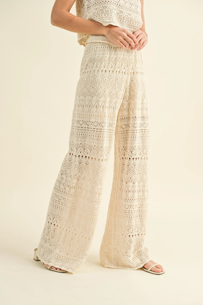 A person is dressed in the MIOU MUSE's Crochet Pattern Pants, a cream-colored cotton two-piece outfit featuring wide-leg pants and a sleeveless top. They complete the look with open-toed shoes set against a soft beige background.