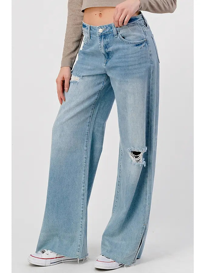 Someone is styling the I&M JEAN, INC. High Rise Skater Jeans in Medium Wash, featuring a chic rip at the right knee. These jeans, with their fashionable hem slit, are ideally paired with a beige cropped sweater and classic white sneakers on a plain white background.