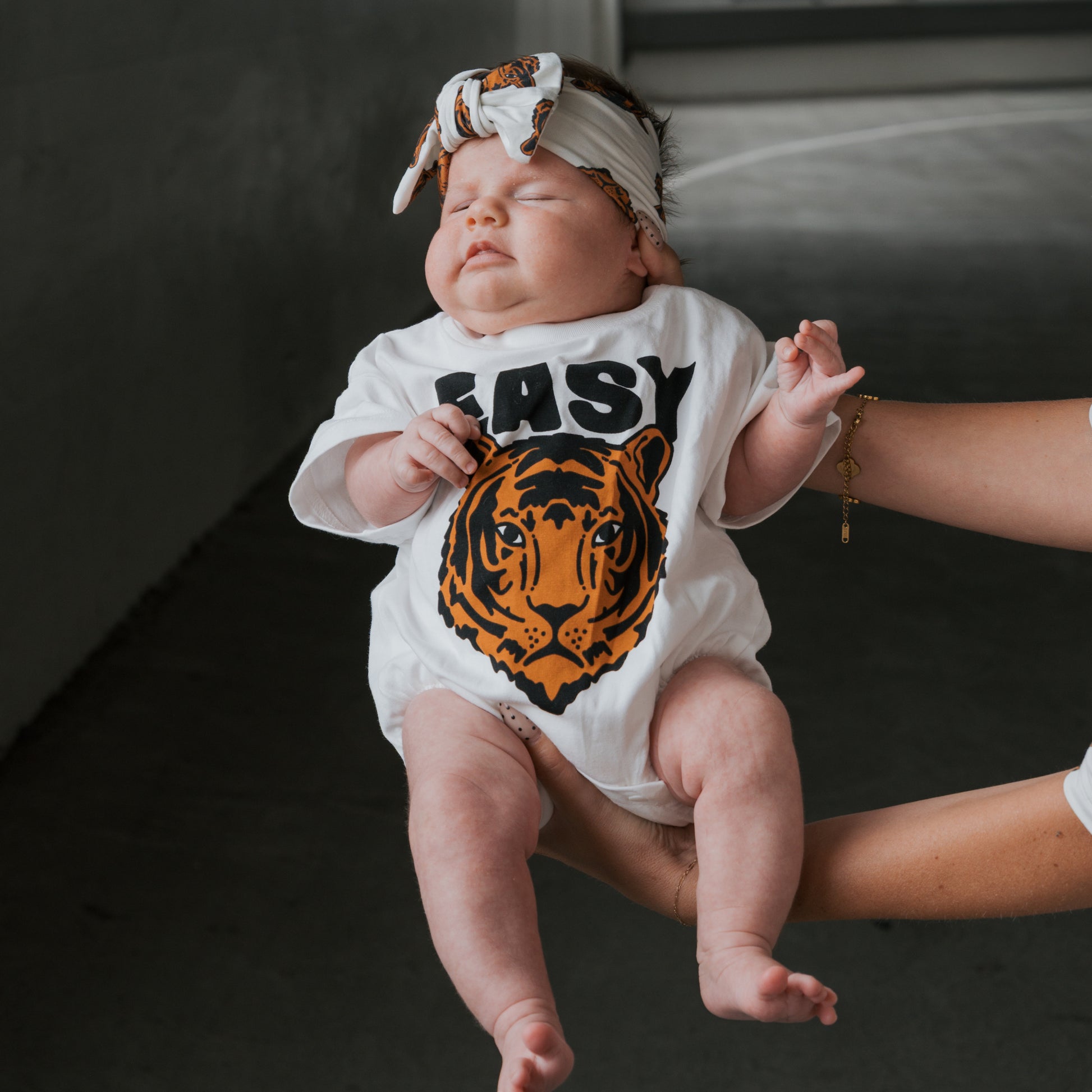 A baby in a white onesie with a tiger face and "EASY" is gently held up, also wearing the forever french baby Bamboo Head Wrap | Tigers—an adjustable headband topped with a tiger print bow for extra style.