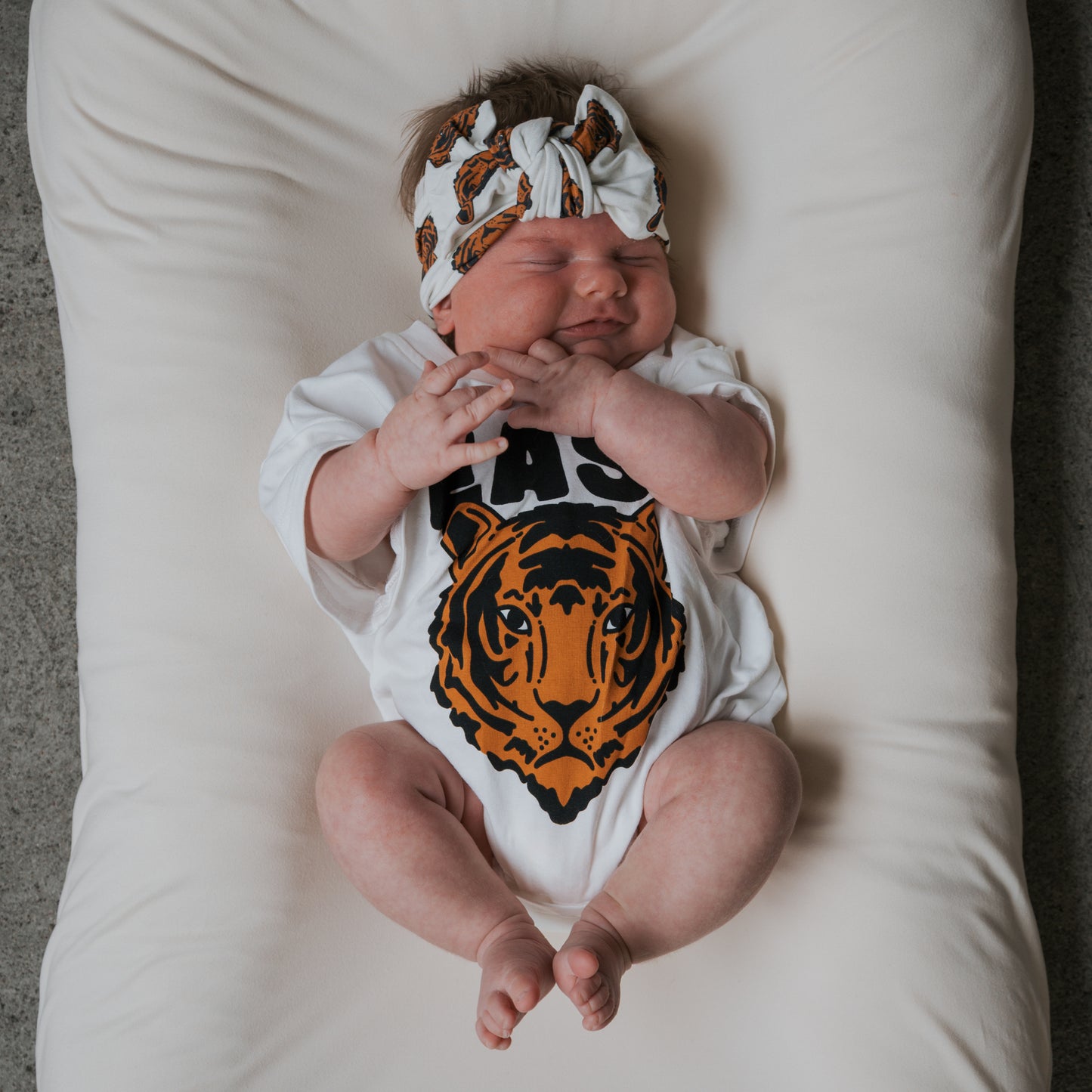 A baby wearing the forever french baby Bamboo Head Wrap | Tigers and a white onesie with a tiger face graphic smiles with eyes closed, lying on a soft white cushion.