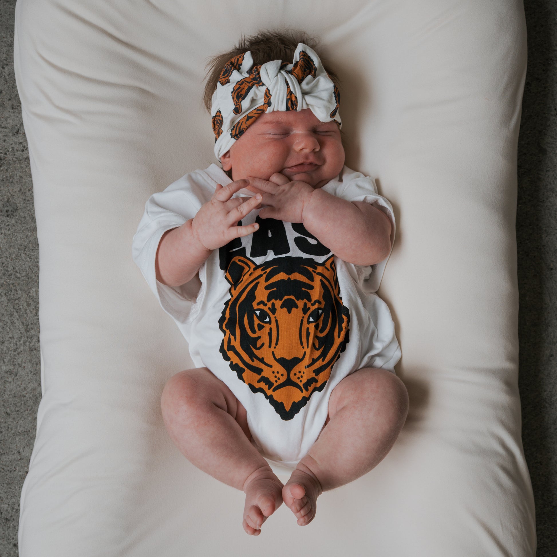 A baby wearing the forever french baby Bamboo Head Wrap | Tigers and a white onesie with a tiger face graphic smiles with eyes closed, lying on a soft white cushion.