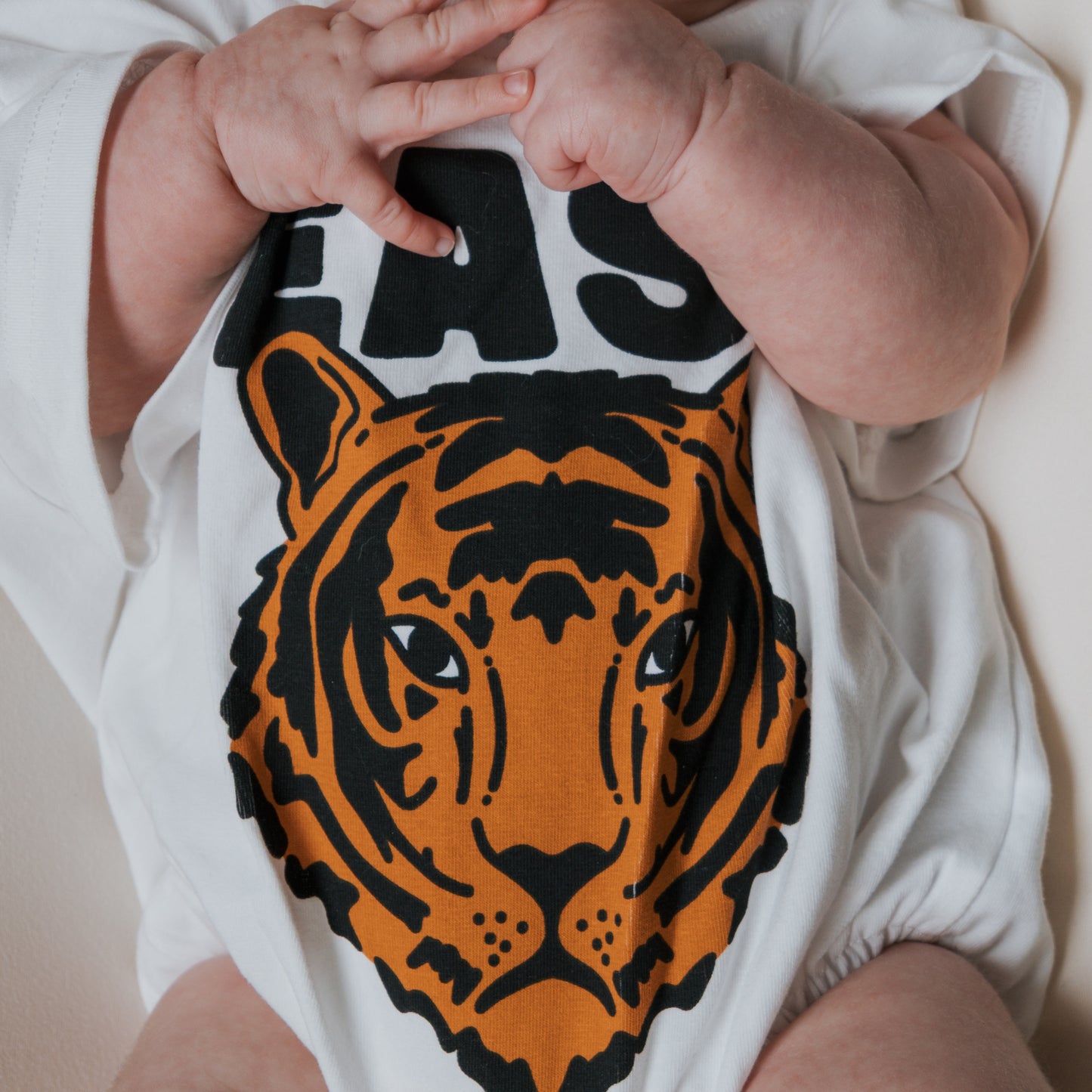 A baby in the forever french baby "Easy Tiger" short sleeve romper, featuring a bold orange and black tiger face graphic, holds their hands together near their mouth.
