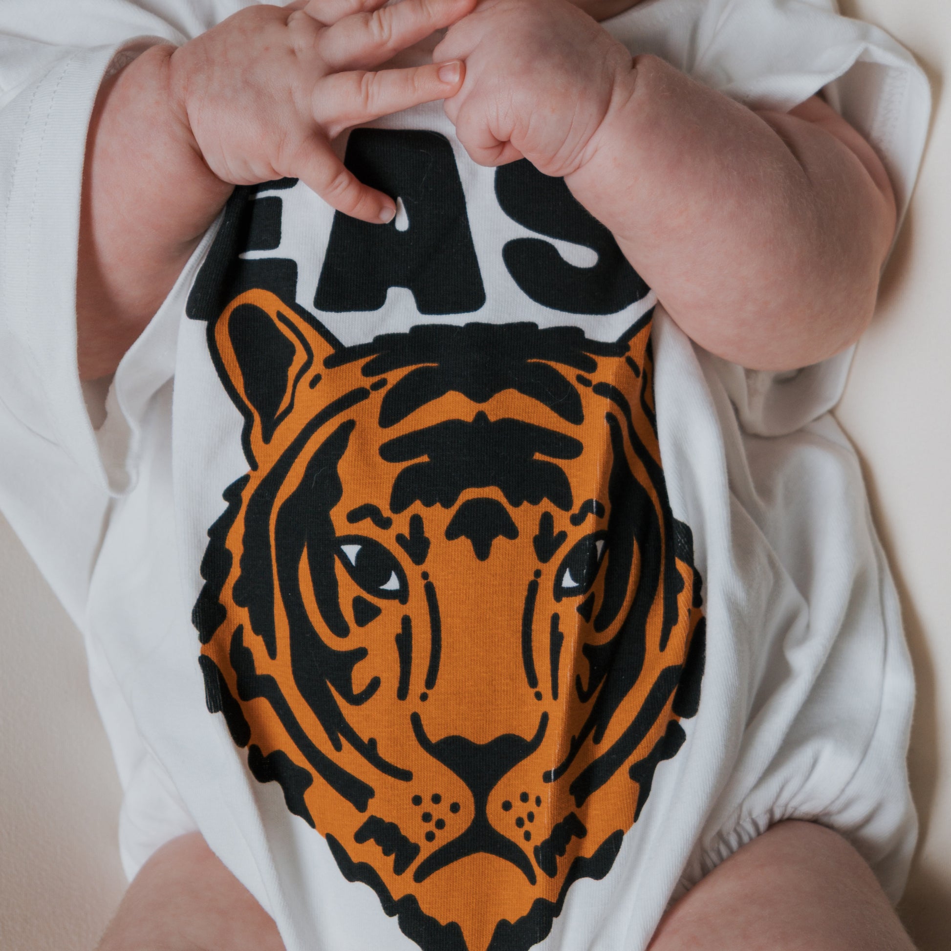 A baby in the forever french baby "Easy Tiger" short sleeve romper, featuring a bold orange and black tiger face graphic, holds their hands together near their mouth.