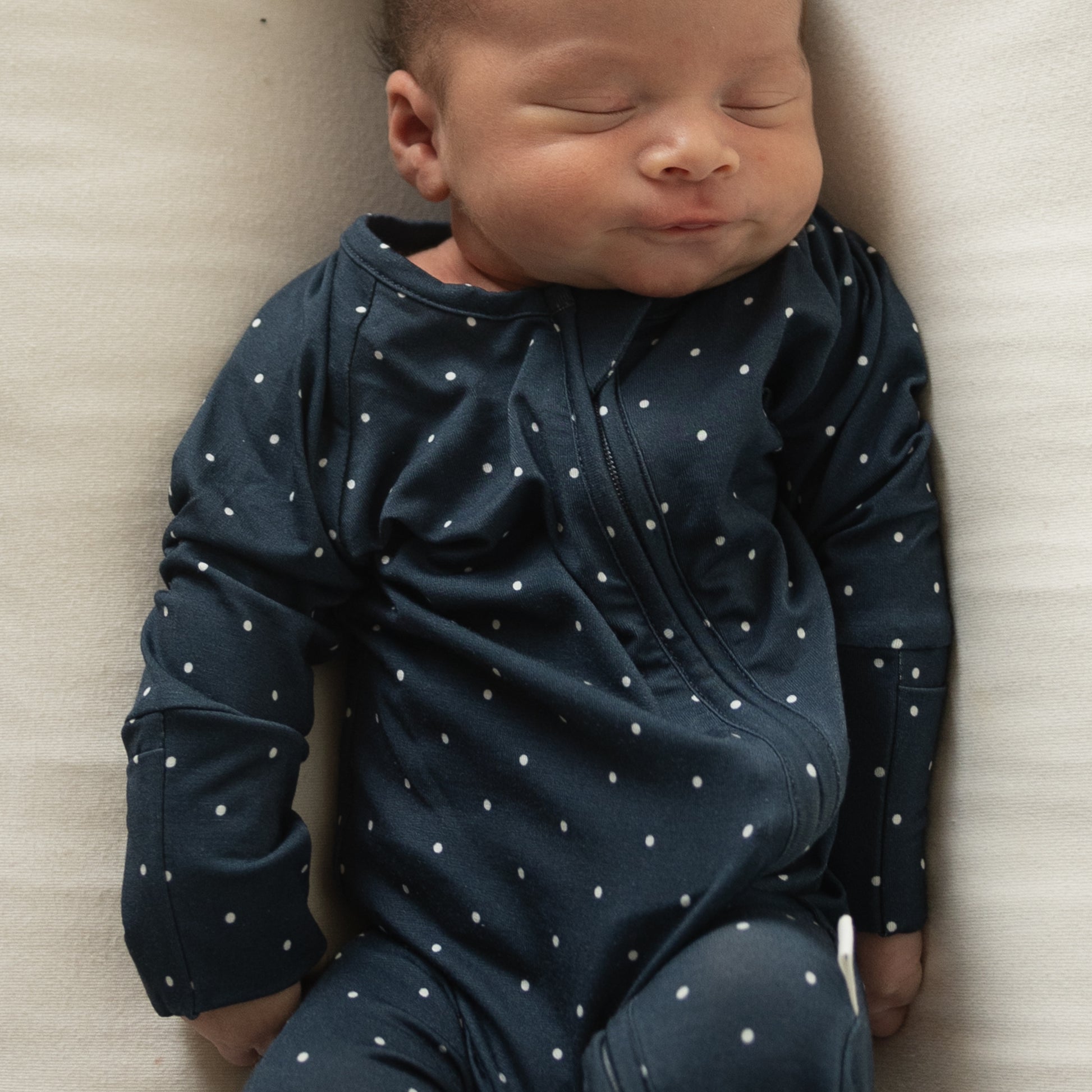 A baby in forever french baby Bamboo Zip Pajamas | Emery—a navy blue onesie with white polka dots—sleeps peacefully on a light blanket, eyes closed and lips slightly pursed.
