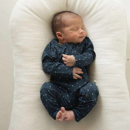 A newborn sleeps on a soft, white cushion, wearing forever french baby Bamboo Zip Pajamas | Emery—a dark blue onesie with white polka dots. Their arms are crossed over the chest and legs are curled up.