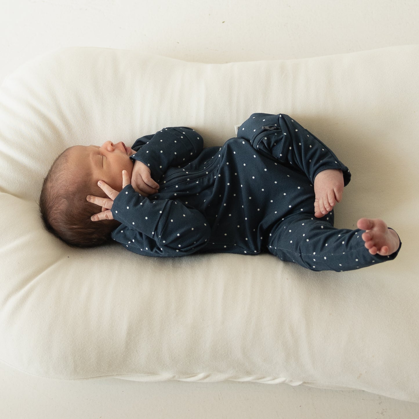 A newborn, dressed in forever french baby’s Bamboo Zip Pajamas | Emery, lies on a soft white cushion with one hand near their face and legs slightly bent, showing off the breathable, hypo-allergenic fabric.