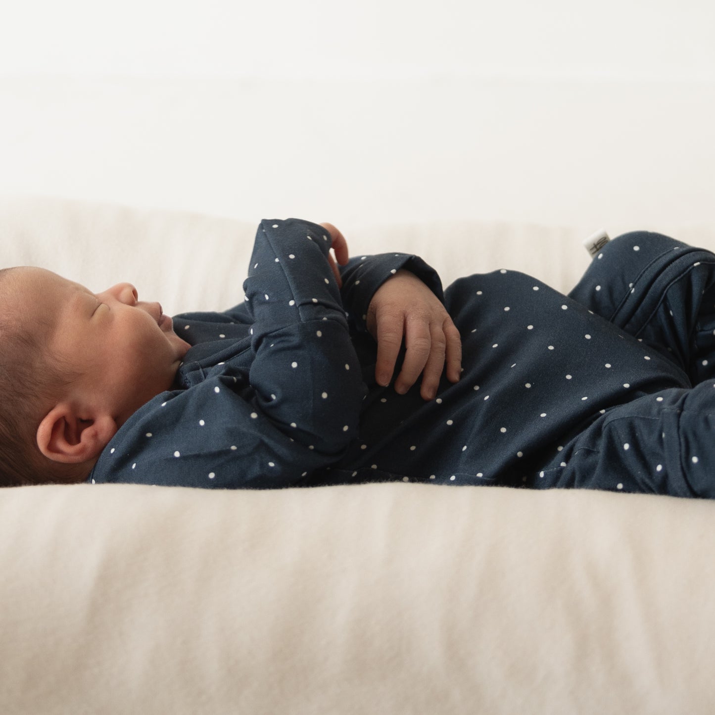 A newborn sleeps on a light blanket, wearing forever french baby’s Bamboo Zip Pajamas | Emery—a navy blue onesie with white polka dots—lying on their back with arms resting on their chest.