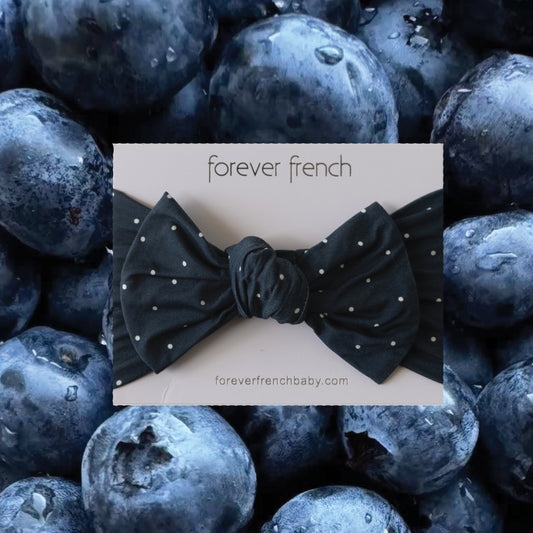 The Bamboo Head Wrap | Emery from forever french baby, a dark blue bow with white polka dots, is displayed on a background of close-up blueberries. The website foreverfrenchbaby.com appears below the bow.