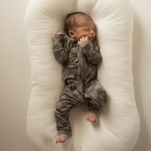 A newborn sleeps on a soft white surface, dressed in forever french baby’s Bamboo Zip Pajamas in Faded Brown Checkerboard—a breathable zip-up onesie. The baby's eyes are closed with one hand near their face.