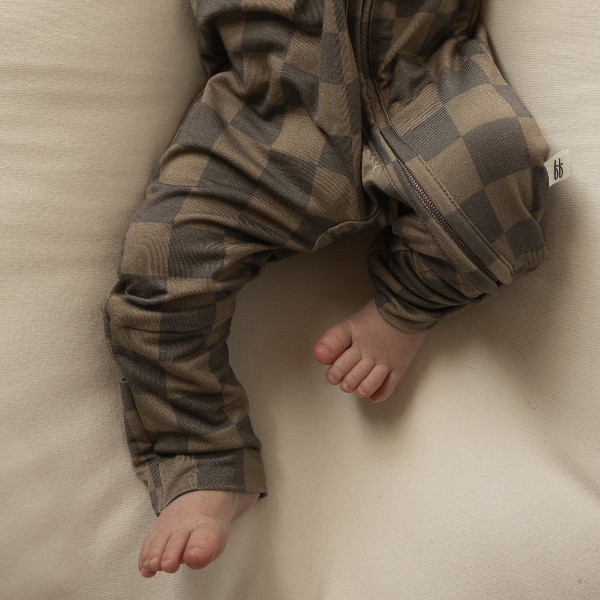 A baby in forever french baby's Bamboo Zip Pajamas in Faded Brown Checkerboard lies on a soft cream surface, their bare feet and legs visible.