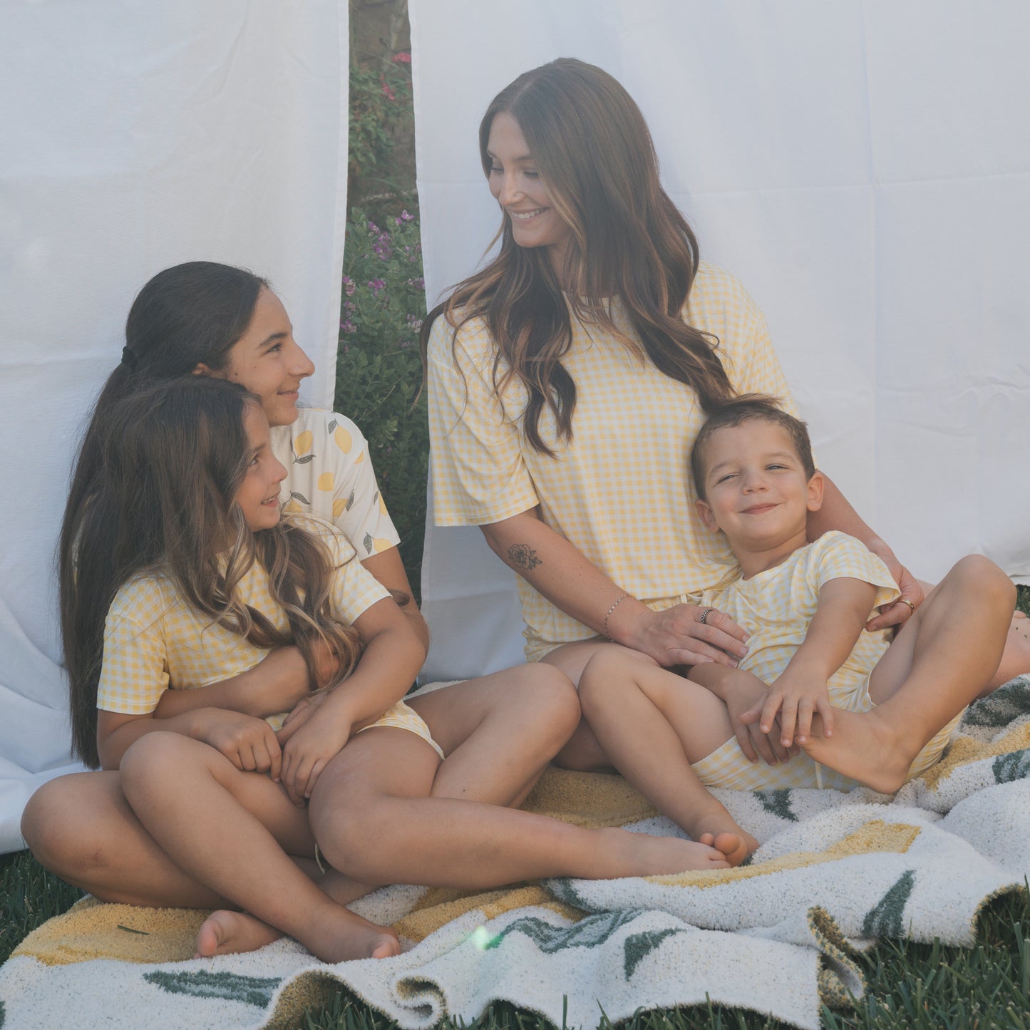 A woman sits outdoors on a blanket with three children, all wearing forever french baby’s Bamboo Short Set in Citron. They smile together, surrounded by green grass and framed by a white sheet behind them.