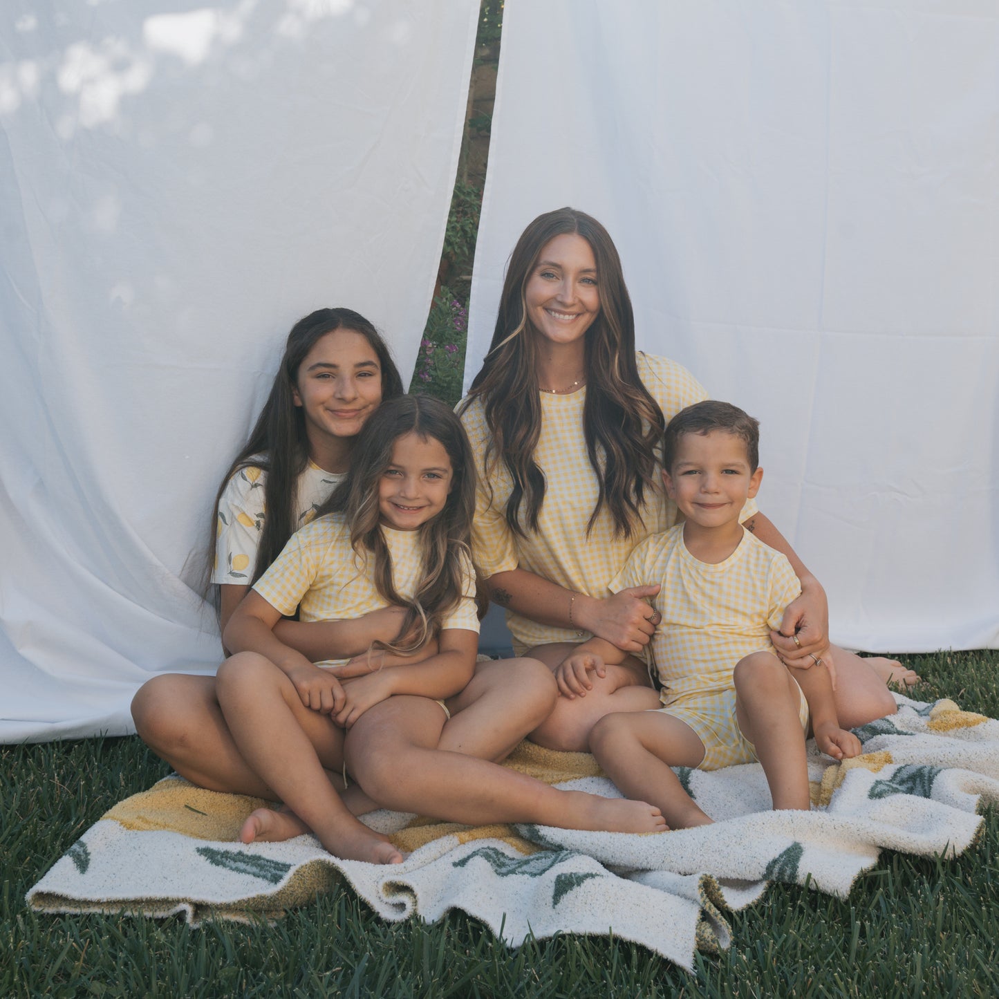 A woman and three children sit together on a blanket outdoors, smiling in matching forever french baby Bamboo Short Set | Citron sleepwear, with a white sheet backdrop and grass beneath them.