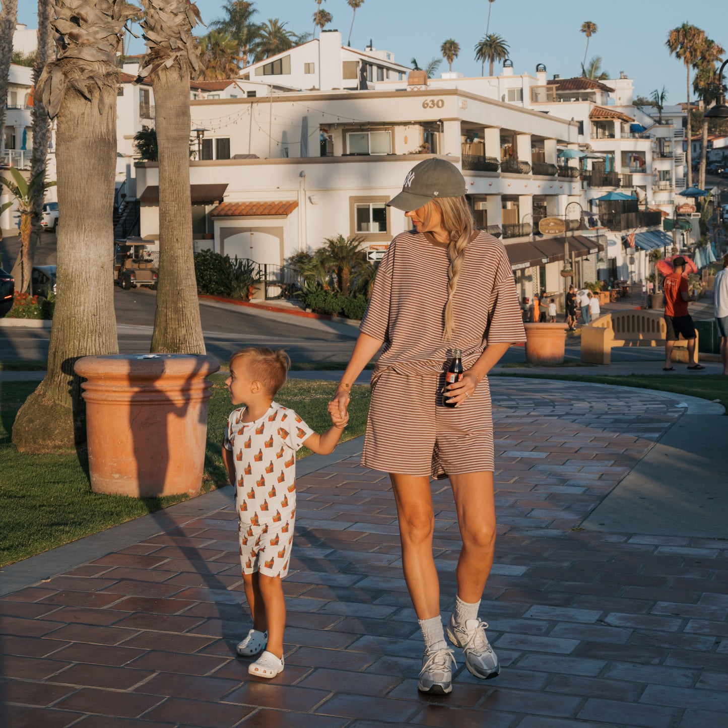 A woman and a young child wearing the Bamboo Short Set | Fries & Cola by forever french baby hold hands on a sunny brick path with palm trees, white buildings, and people nearby. Their soft, breathable outfits ensure all-day comfort.