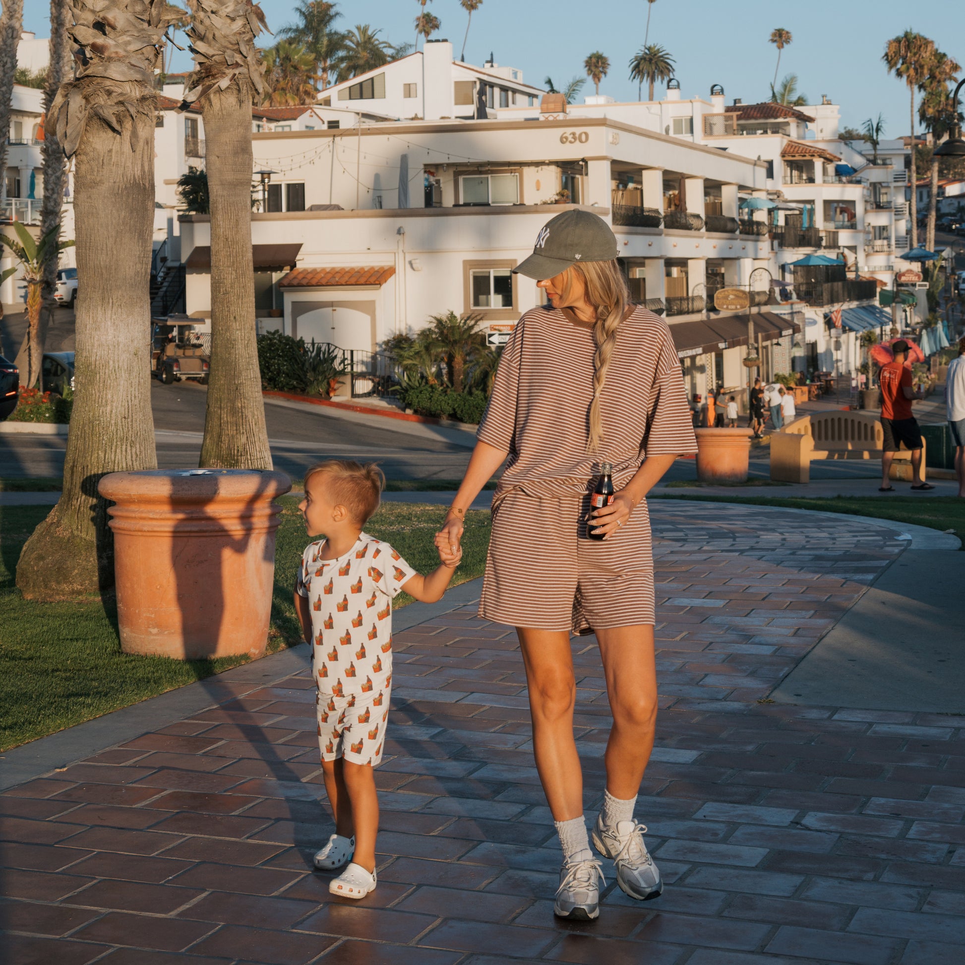 A woman and a young child wearing the Bamboo Short Set | Fries & Cola by forever french baby hold hands on a sunny brick path with palm trees, white buildings, and people nearby. Their soft, breathable outfits ensure all-day comfort.
