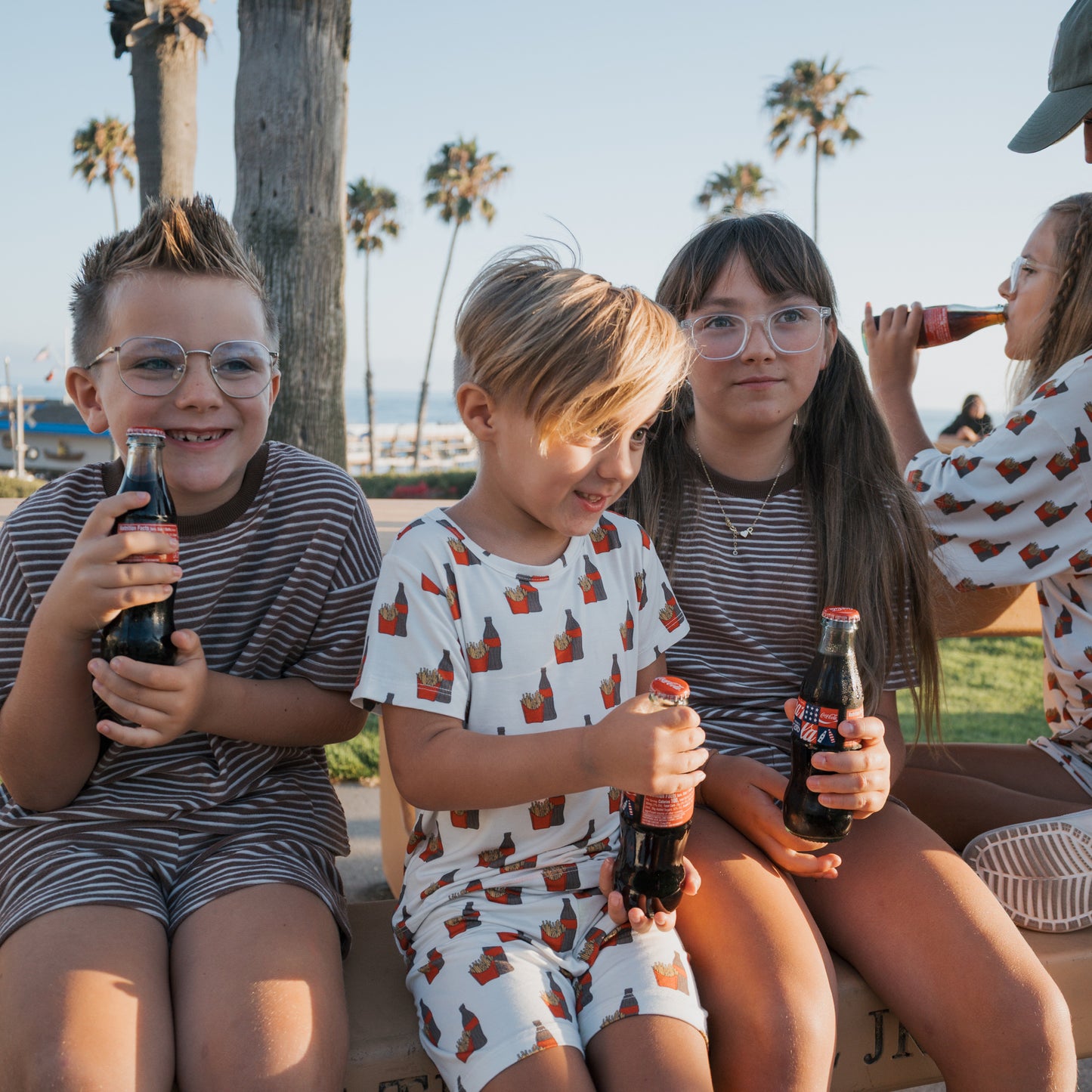 Three children wearing forever french baby Child Terry Short Set | Cola Stripe sit on a bench holding glass Coca-Cola bottles. Palm trees, the beach, and a family in matching outfits enjoying drinks are in the background.