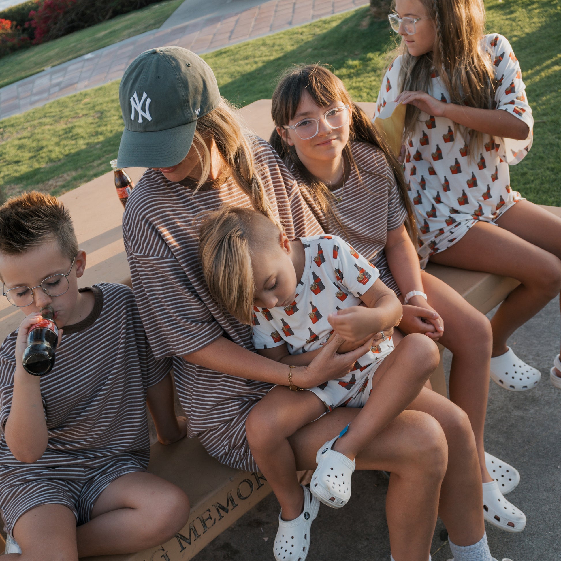 A woman and four children sit on a bench outdoors in matching forever french baby Bamboo Short Sets | Fries & Cola. One child drinks from a bottle, another rests their head on her lap. The vibe is relaxed and casual.
