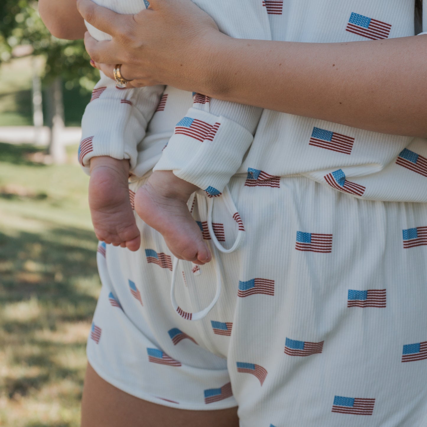 An adult holds a baby outdoors, both wearing matching forever french baby Bamboo Zip Pajamas | Americana Ribbed. Their torsos, arms, and the baby’s bare feet are visible against a blurred background of grass and trees.