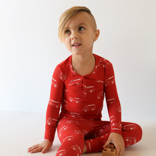 A young blonde child sits on the floor in forever french baby's Bamboo Two Piece Pajamas | Fire Truck, holding wooden blocks and gazing up curiously. The red, breathable pajamas with white fire truck patterns pop against a plain white background.