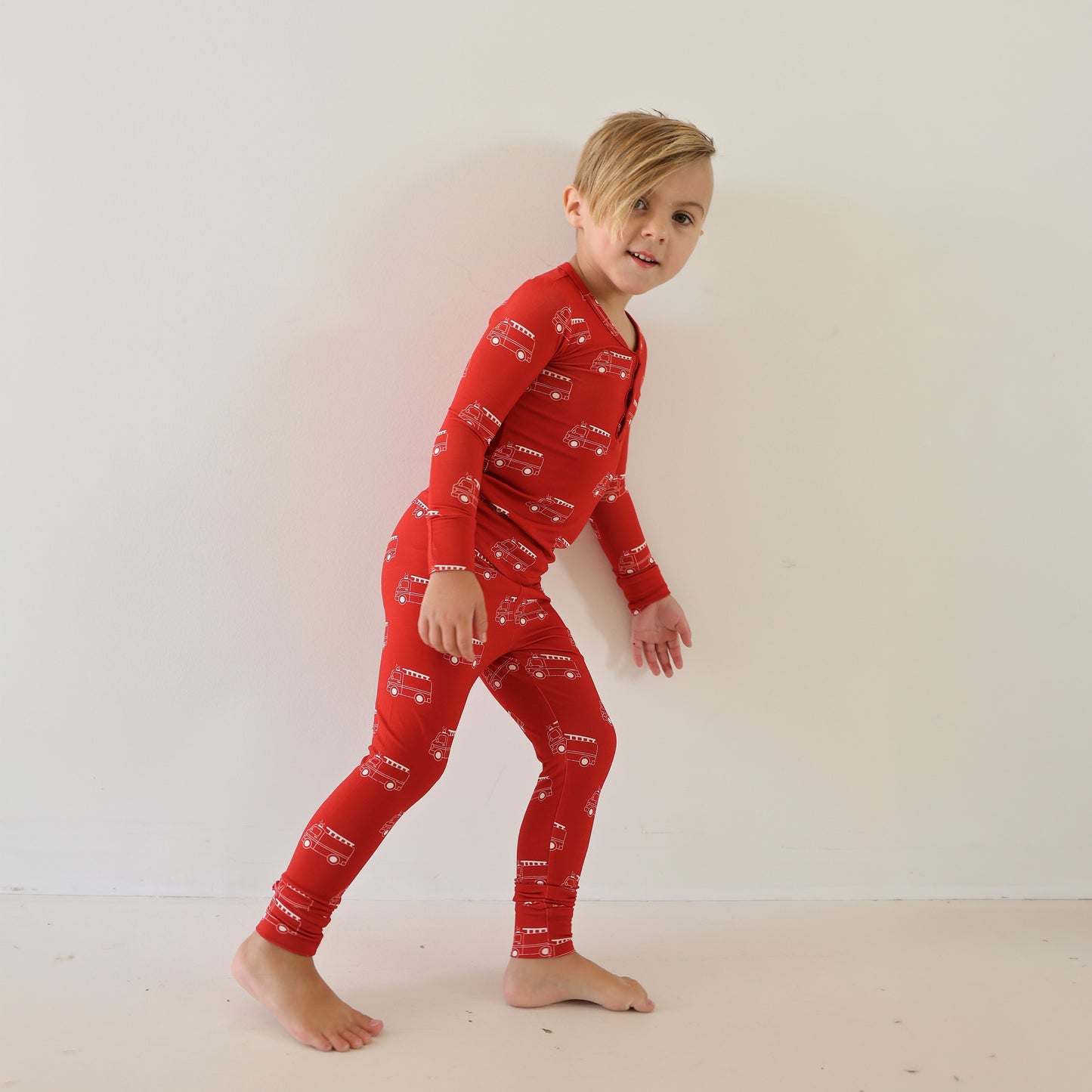 A young child with blonde hair smiles at the camera, wearing forever french baby’s Bamboo Two Piece Pajamas | Fire Truck—a red pajama set with white fire truck patterns—standing barefoot against a plain white wall.