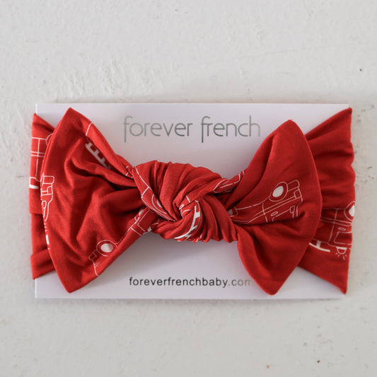 The Bamboo Head Wrap | Fire Truck by forever french baby is a red knot headband with white fire truck print, shown on a branded display card on a light-colored surface.
