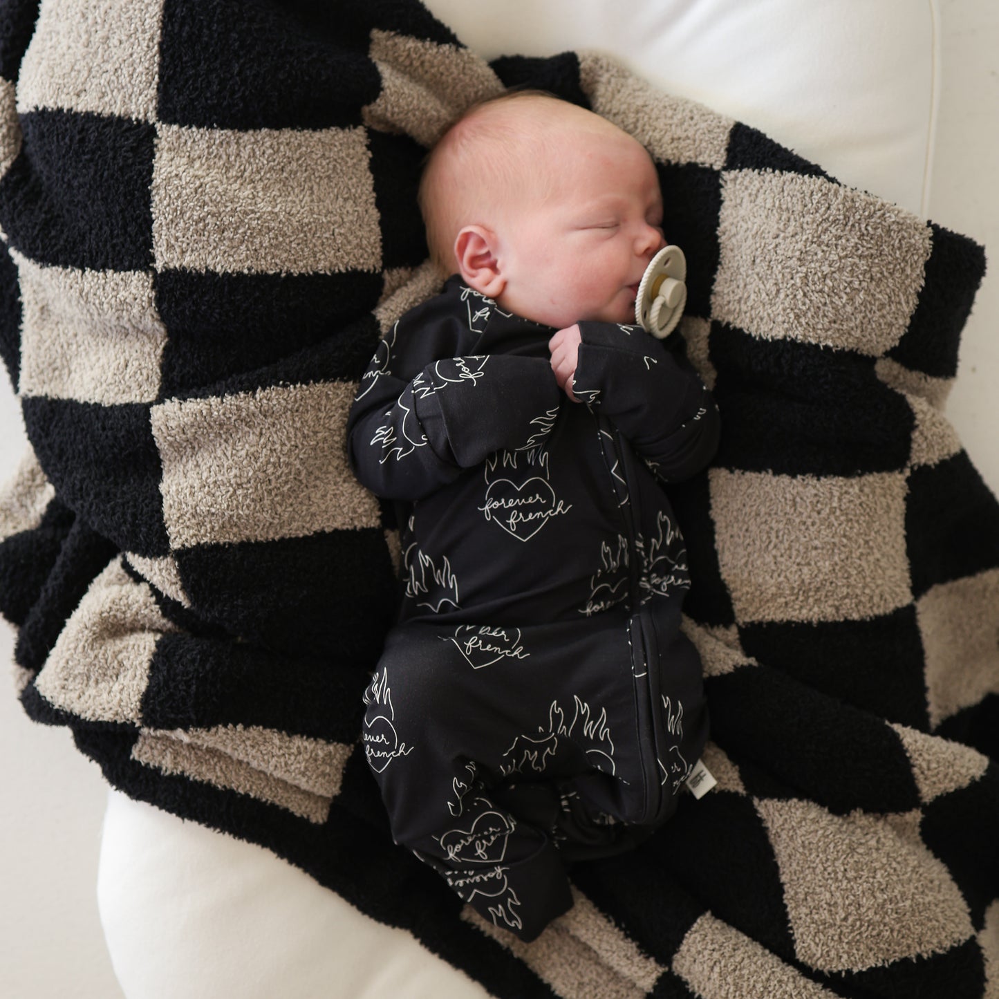 A newborn sleeps with a pacifier, wrapped in a black and beige checkered blanket, wearing forever french baby Bamboo Zip Pajamas | Flaming Hearts—soft and breathable baby sleepwear.