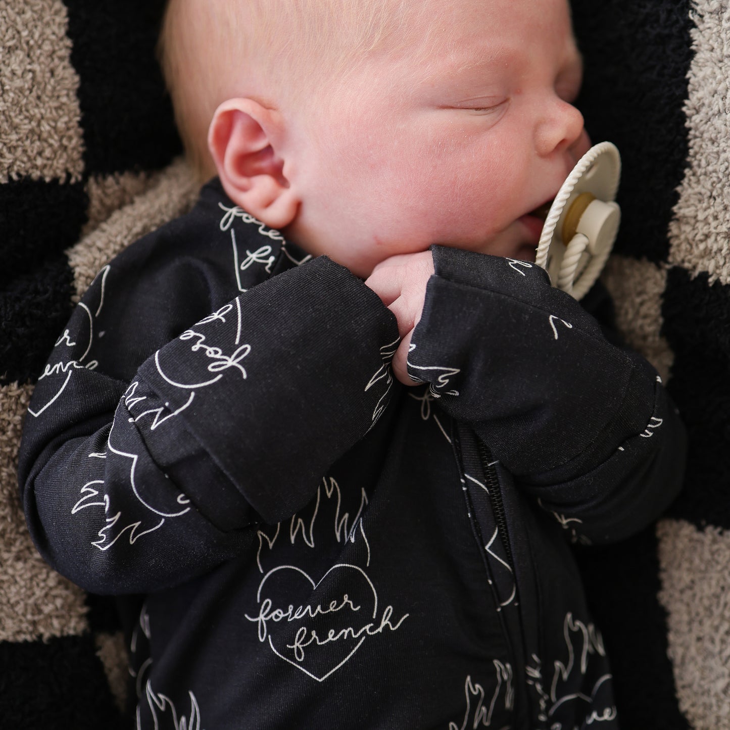 A baby in forever french baby's Bamboo Zip Pajamas | Flaming Hearts—a black onesie with white heart and flame designs—sleeps with a beige pacifier on a black and beige checkered blanket.