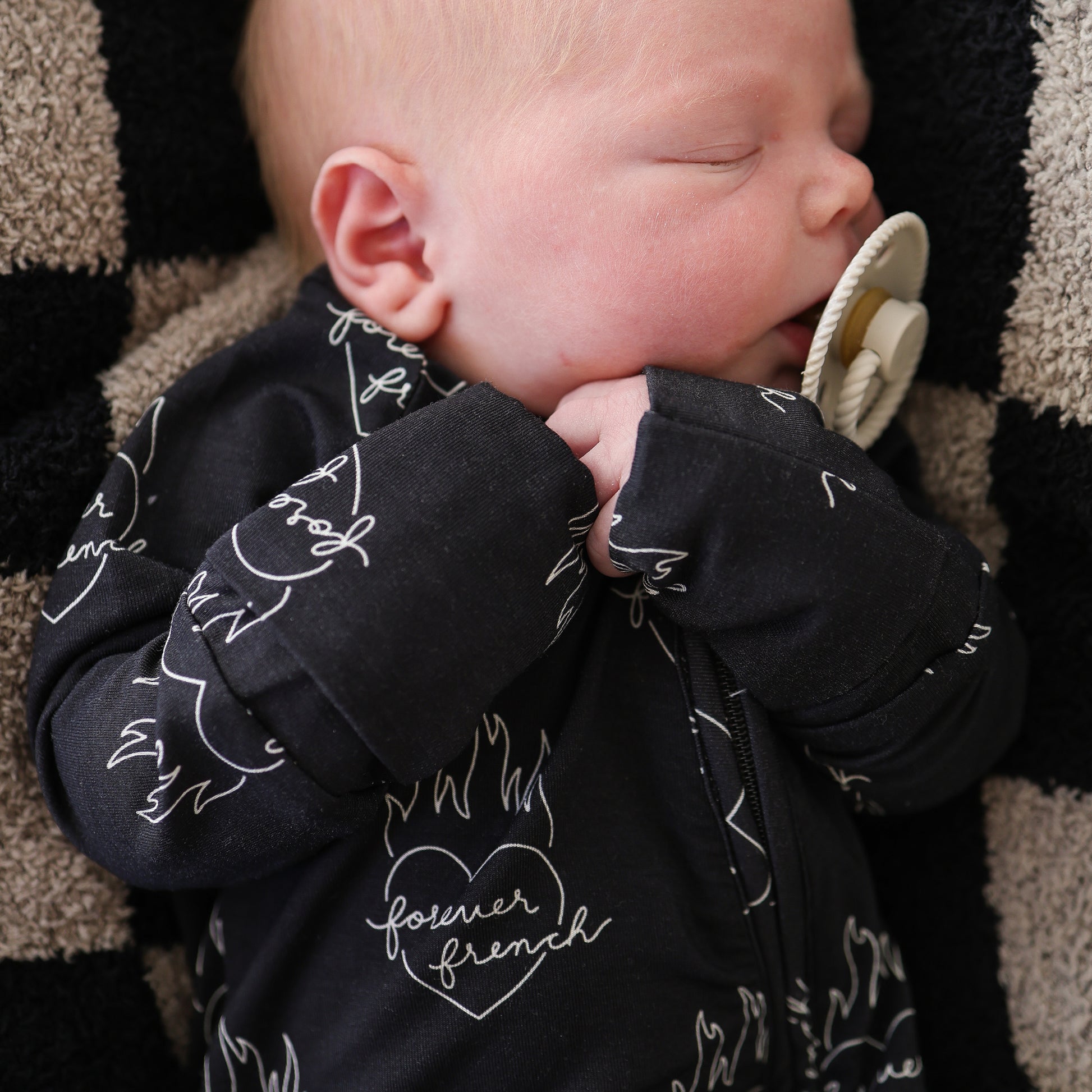 A baby in forever french baby's Bamboo Zip Pajamas | Flaming Hearts—a black onesie with white heart and flame designs—sleeps with a beige pacifier on a black and beige checkered blanket.