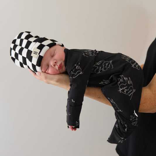 A newborn wearing a forever french baby ff Knit Beanie in Black + White Checkerboard and a black patterned outfit sleeps peacefully in an adult's arms against a plain background.