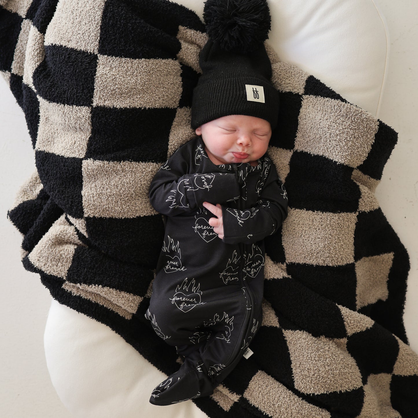 A newborn sleeps on a forever french baby Plush Blanket | Checkmate Checkerboard, wearing a black beanie with pom-pom and a matching black outfit with white designs.
