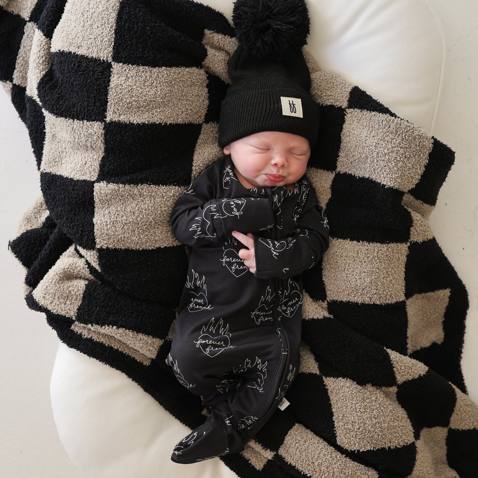A newborn sleeps on a forever french baby Plush Blanket | Checkmate Checkerboard, wearing a black beanie with pom-pom and a matching black outfit with white designs.