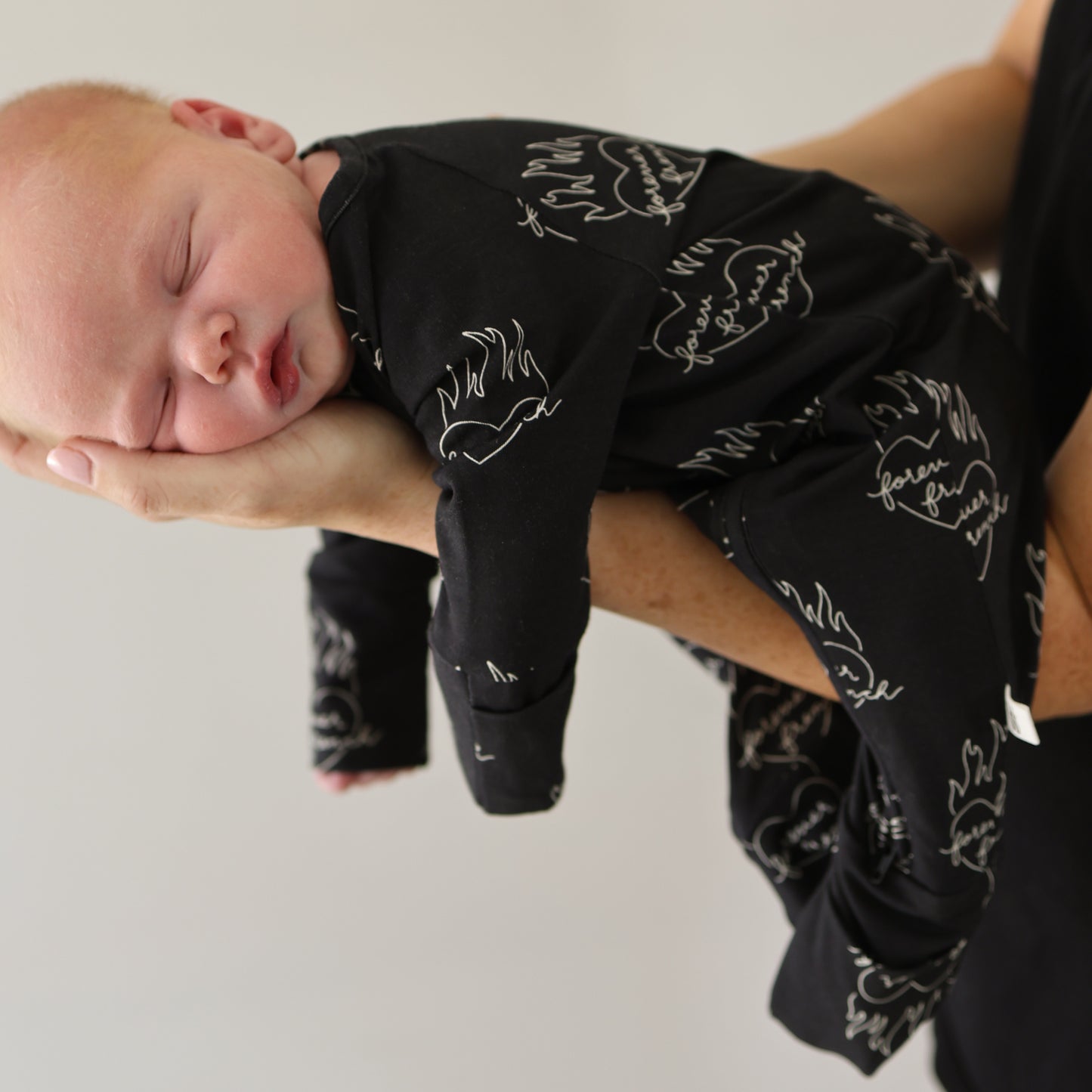 A newborn sleeps peacefully in forever french baby's Bamboo Zip Pajamas | Flaming Hearts, nestled gently in an adult's hand against a neutral background.