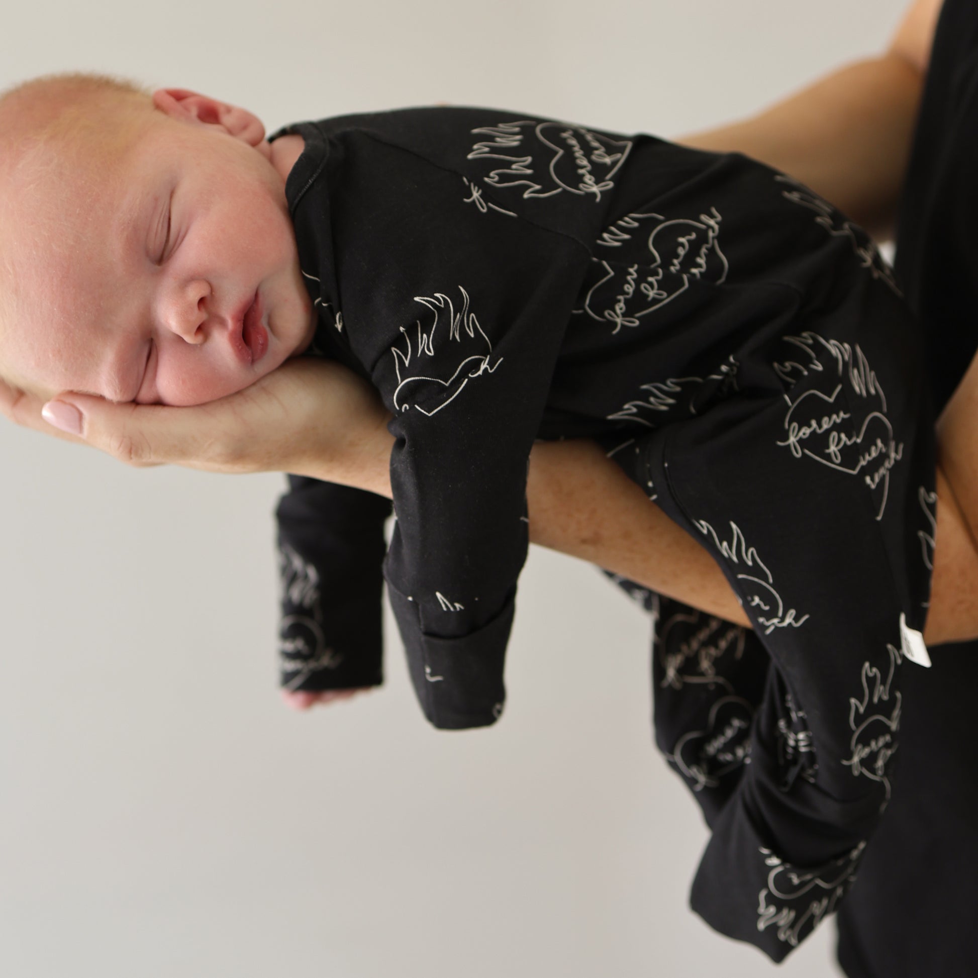 A newborn sleeps peacefully in forever french baby's Bamboo Zip Pajamas | Flaming Hearts, nestled gently in an adult's hand against a neutral background.