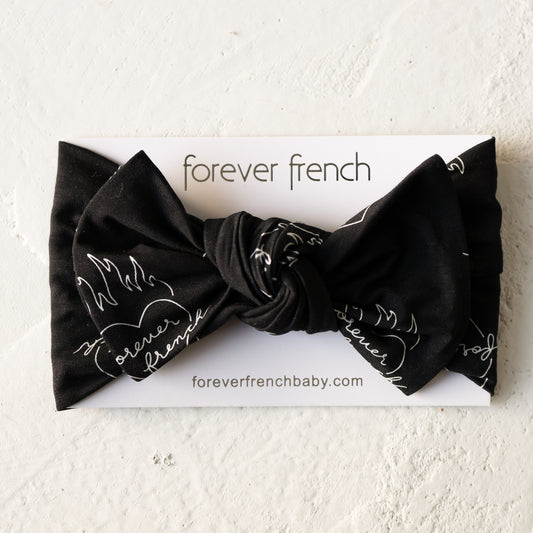 The Bamboo Head Wrap | Flaming Hearts by forever french baby features a black fabric bow with white script and flame design, displayed on branded packaging atop a light textured surface.
