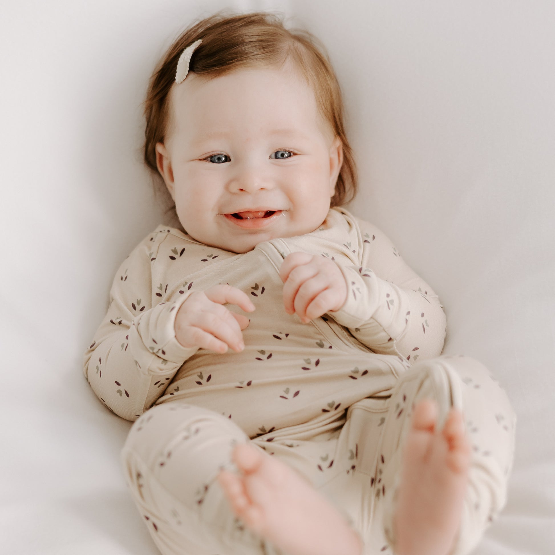 A smiling light-skinned baby with light brown hair wears forever french baby Bamboo Zip Pajamas | Flora, a cream onesie with a small leaf pattern, and lies on a white surface with feet up and hands near the mouth.
