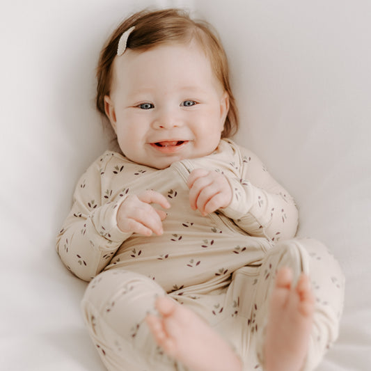 A smiling light-skinned baby with light brown hair wears forever french baby Bamboo Zip Pajamas | Flora, a cream onesie with a small leaf pattern, and lies on a white surface with feet up and hands near the mouth.