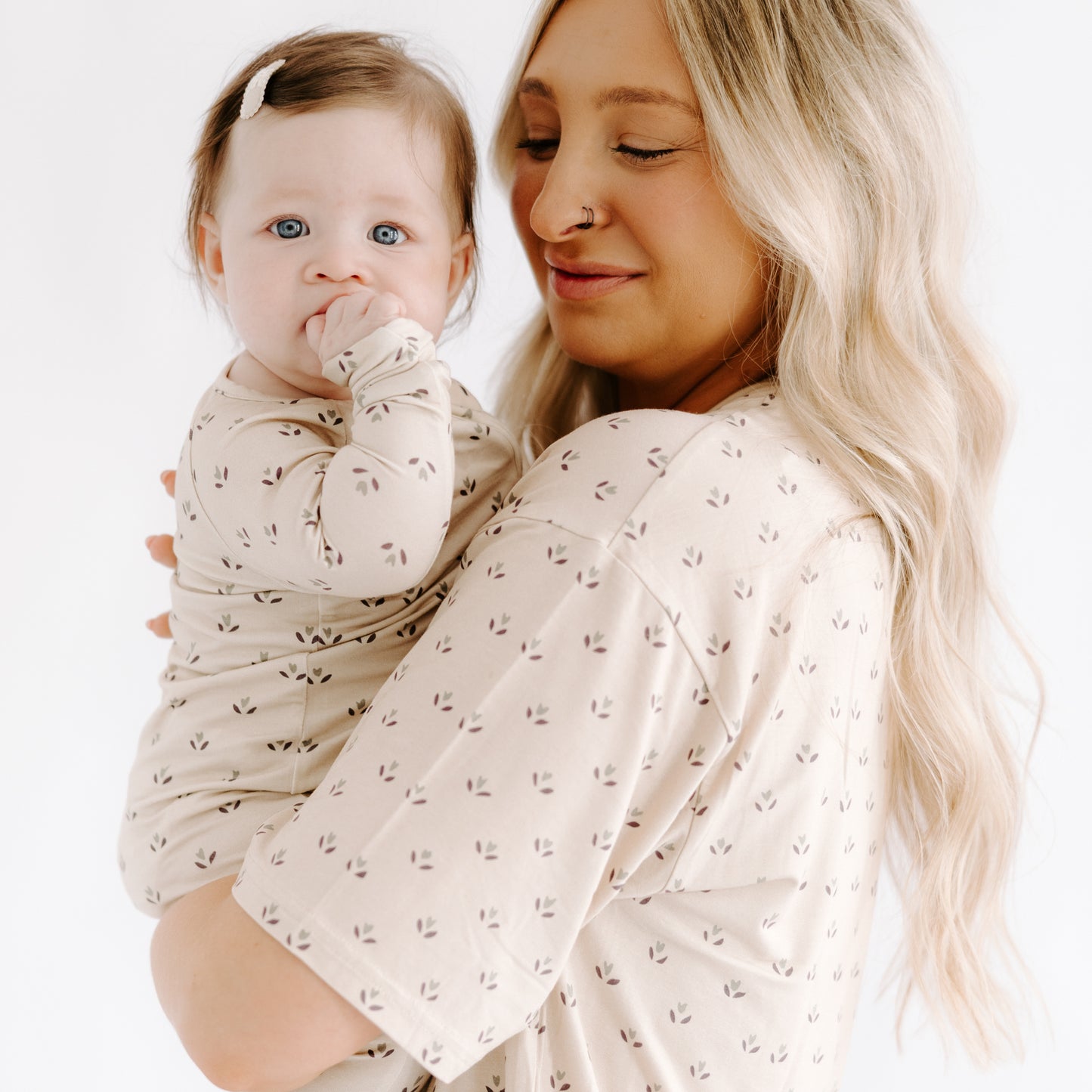A woman with long blonde hair holds a baby wearing Forever French Baby's Bamboo Zip Pajamas in Flora. Both wear matching cream outfits with small leaf patterns, facing the camera against a white background.