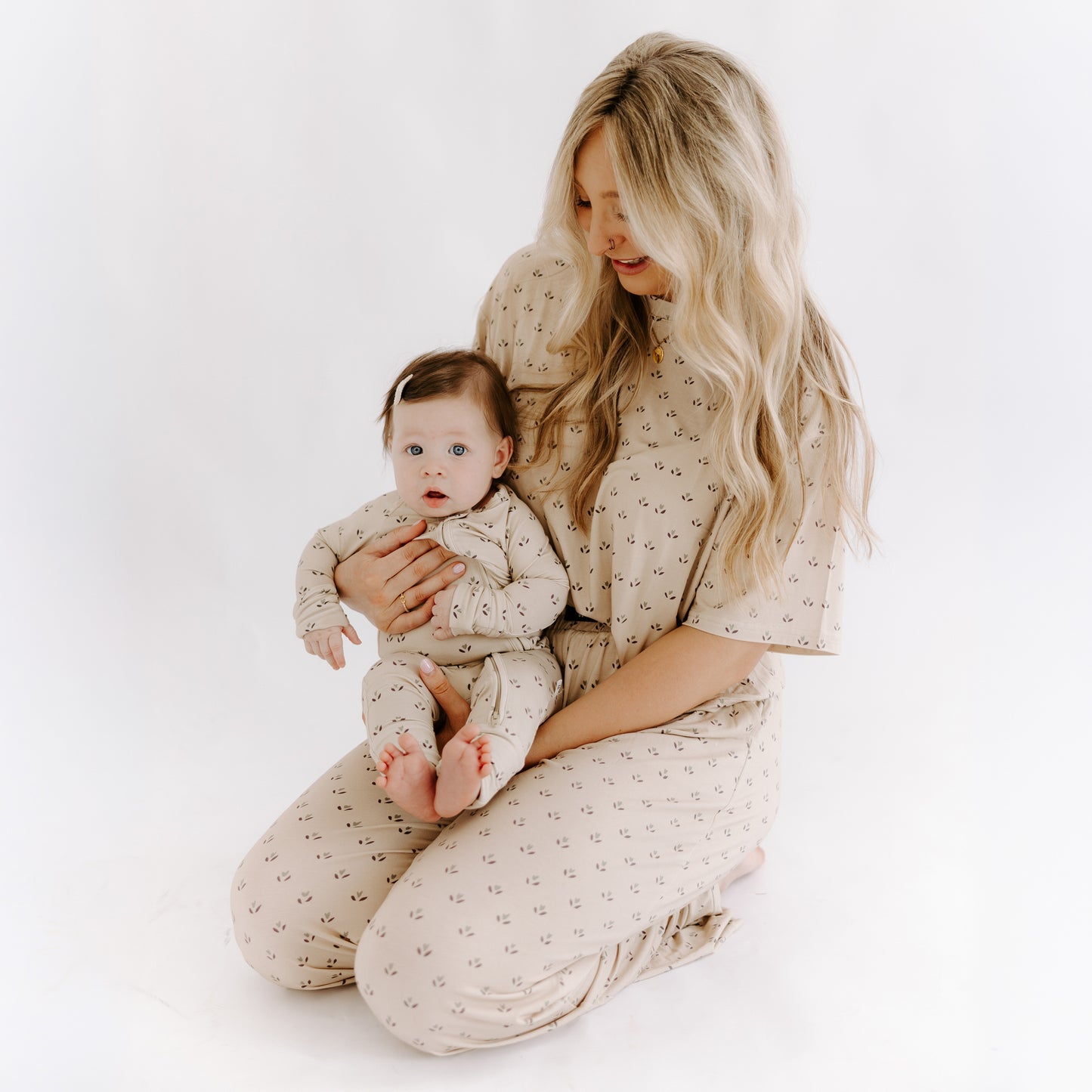 A woman with long blonde hair kneels, holding a baby in her lap. Both wear matching forever french baby Bamboo Zip Pajamas | Flora—beige with a small floral print—ideal breathable sleepwear, set against a white minimal background.