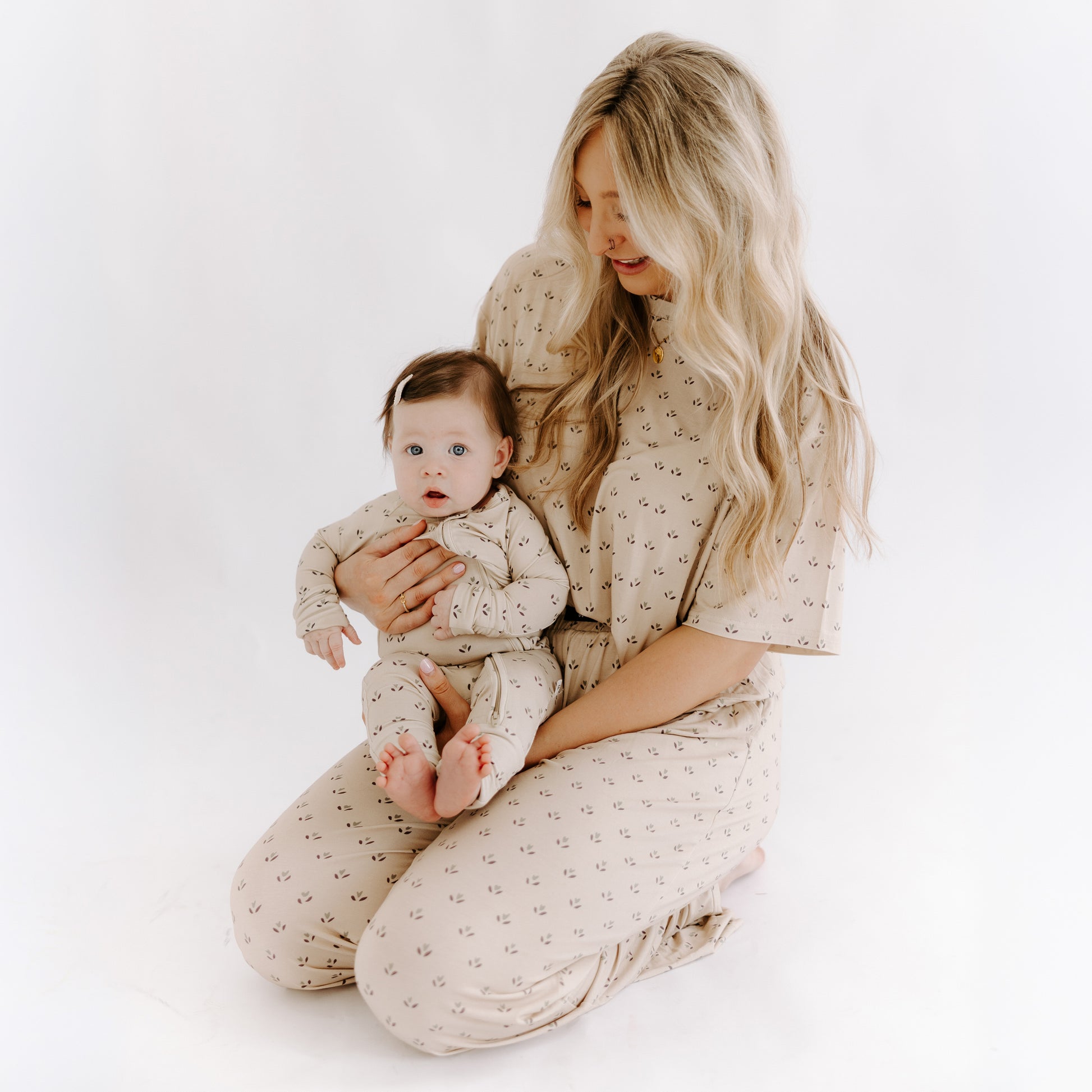 A woman with long blonde hair kneels, holding a baby in her lap. Both wear matching forever french baby Bamboo Zip Pajamas | Flora—beige with a small floral print—ideal breathable sleepwear, set against a white minimal background.