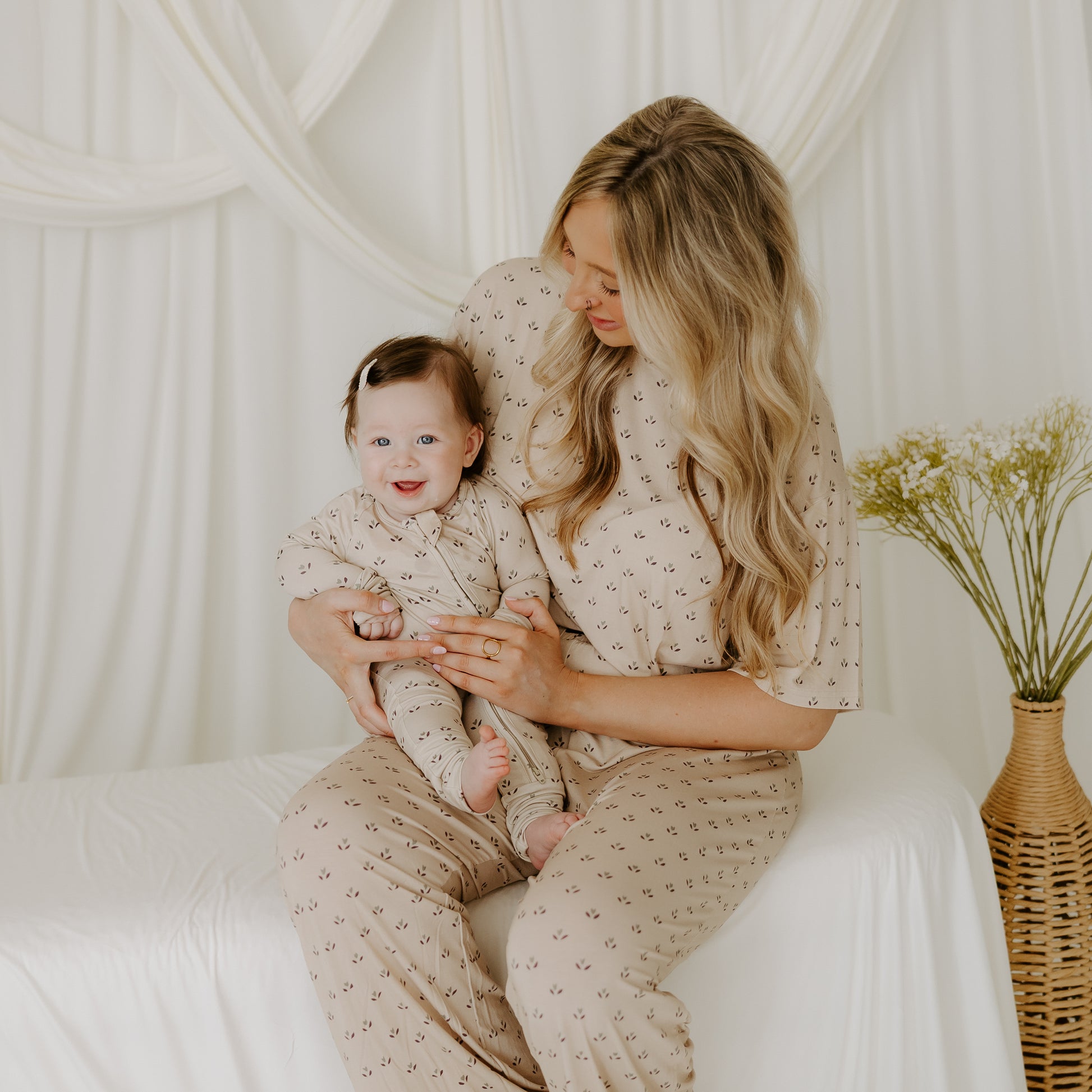 A woman with long blonde hair sits on a white bench holding a smiling baby. Both wear matching forever french baby Bamboo Zip Pajamas | Flora, featuring small patterns. White drapes and yellow flowers are in the background.