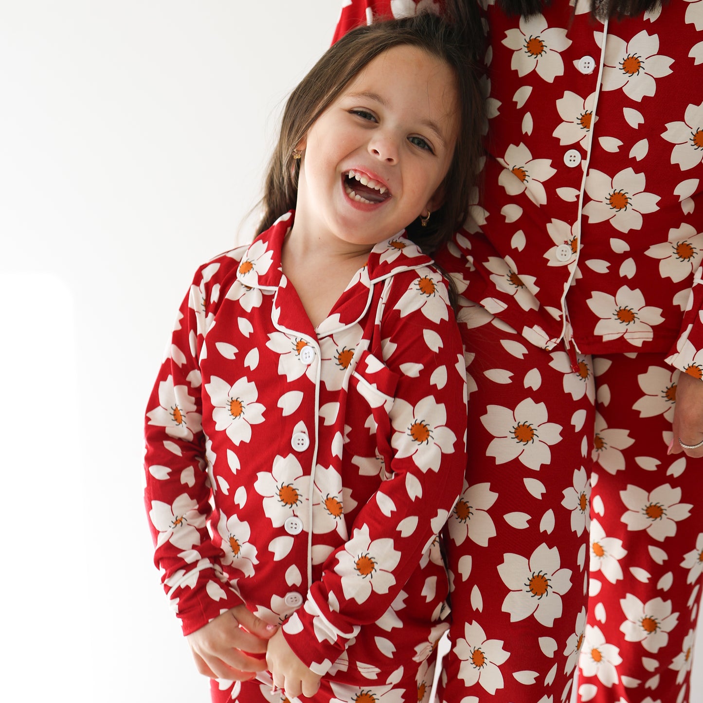 A young child smiles brightly in the forever french baby Girl's Bamboo Button Up Lounge Set | Cherry Blossoms, standing beside an adult in matching pajamas against a plain white background.