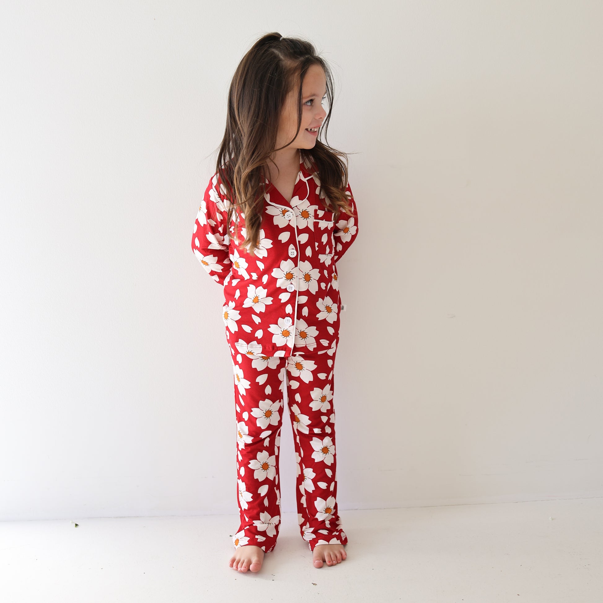 A young girl with long brown hair stands barefoot against a white wall, smiling and looking to the side. She is wearing the forever french baby Girl's Bamboo Button Up Lounge Set in the Cherry Blossoms pattern, with her hands tucked behind her back.