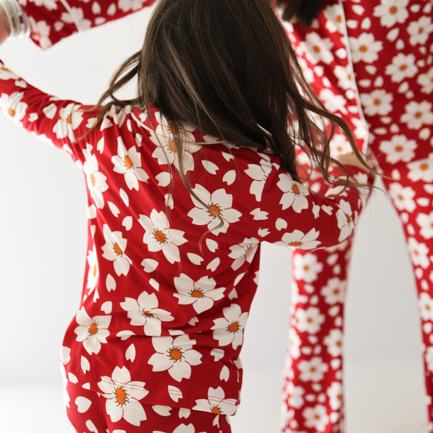 A child wears the forever french baby Girl's Bamboo Button Up Lounge Set | Cherry Blossoms, dancing beside an adult in matching pajamas. Both face away from the camera in a brightly lit room.