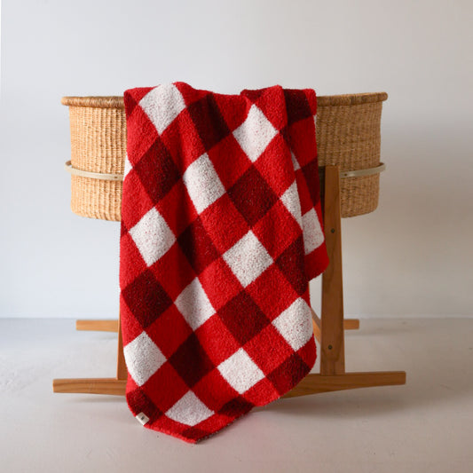 The forever french baby Plush Blanket | Scarlet Gingham, a soft Oeko-Tex certified red and white checkered blanket, is draped over a woven bassinet with wooden legs against a plain white background.